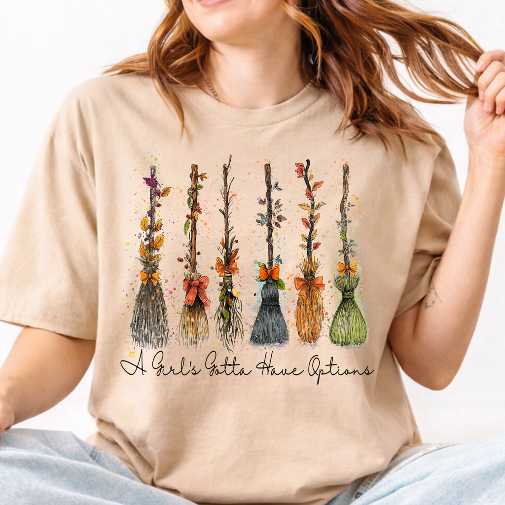 Woman wearing a sand Halloween graphic tee with colorful witch brooms and A Girl’s Gotta Have Options lettering.