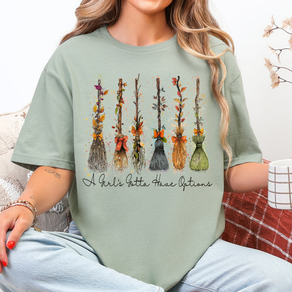 Woman wearing a sage Halloween graphic tee with colorful witch brooms and A Girl’s Gotta Have Options lettering.