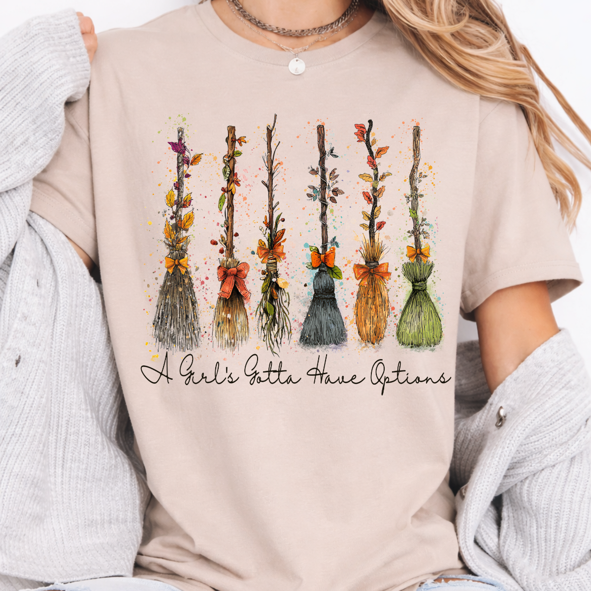 Woman wearing a sand Halloween graphic tee with colorful witch brooms and A Girl’s Gotta Have Options lettering.