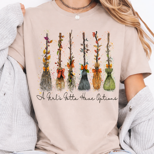 Woman wearing a sand Halloween graphic tee with colorful witch brooms and A Girl’s Gotta Have Options lettering.