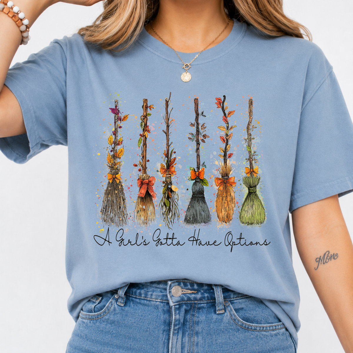 Woman wearing a stone blue Halloween graphic tee with colorful witch brooms and A Girl’s Gotta Have Options lettering.
