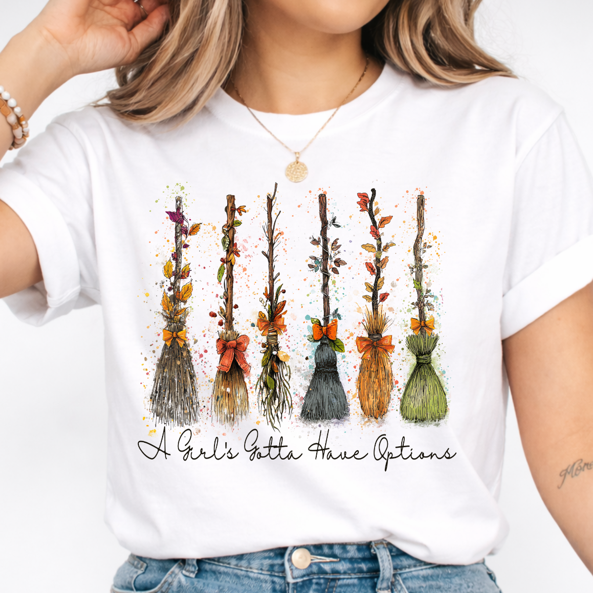 Woman wearing a white Halloween graphic tee with colorful witch brooms and A Girl’s Gotta Have Options lettering.
