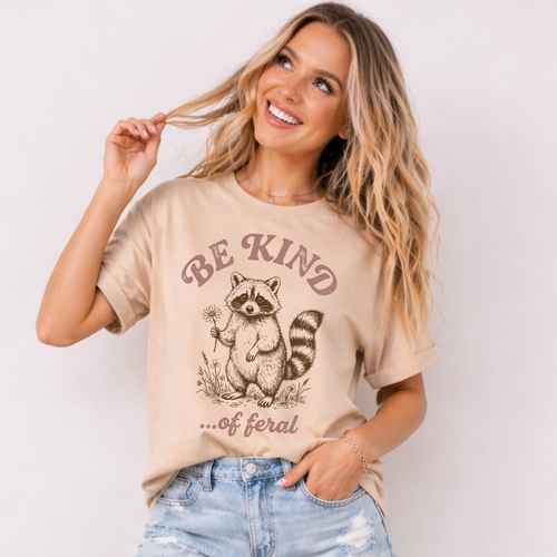 Woman wearing a beige t-shirt with a raccoon graphic and 'Be Kind...of Feral' text on a plain background