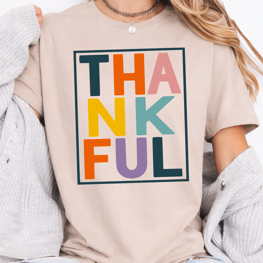 Woman wearing a sand Thankful Thanksgiving graphic tee with colorful block lettering.