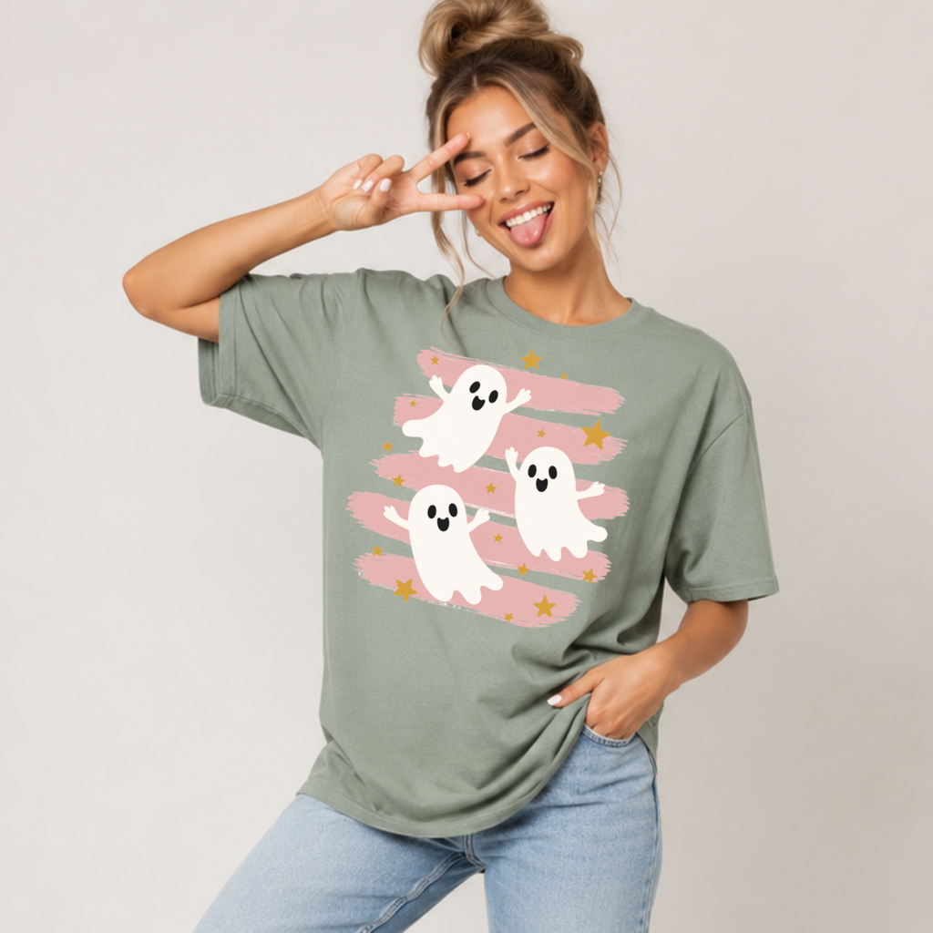 Woman wearing a sage green Halloween graphic tee with three white ghosts, pink brushstrokes, and gold stars.