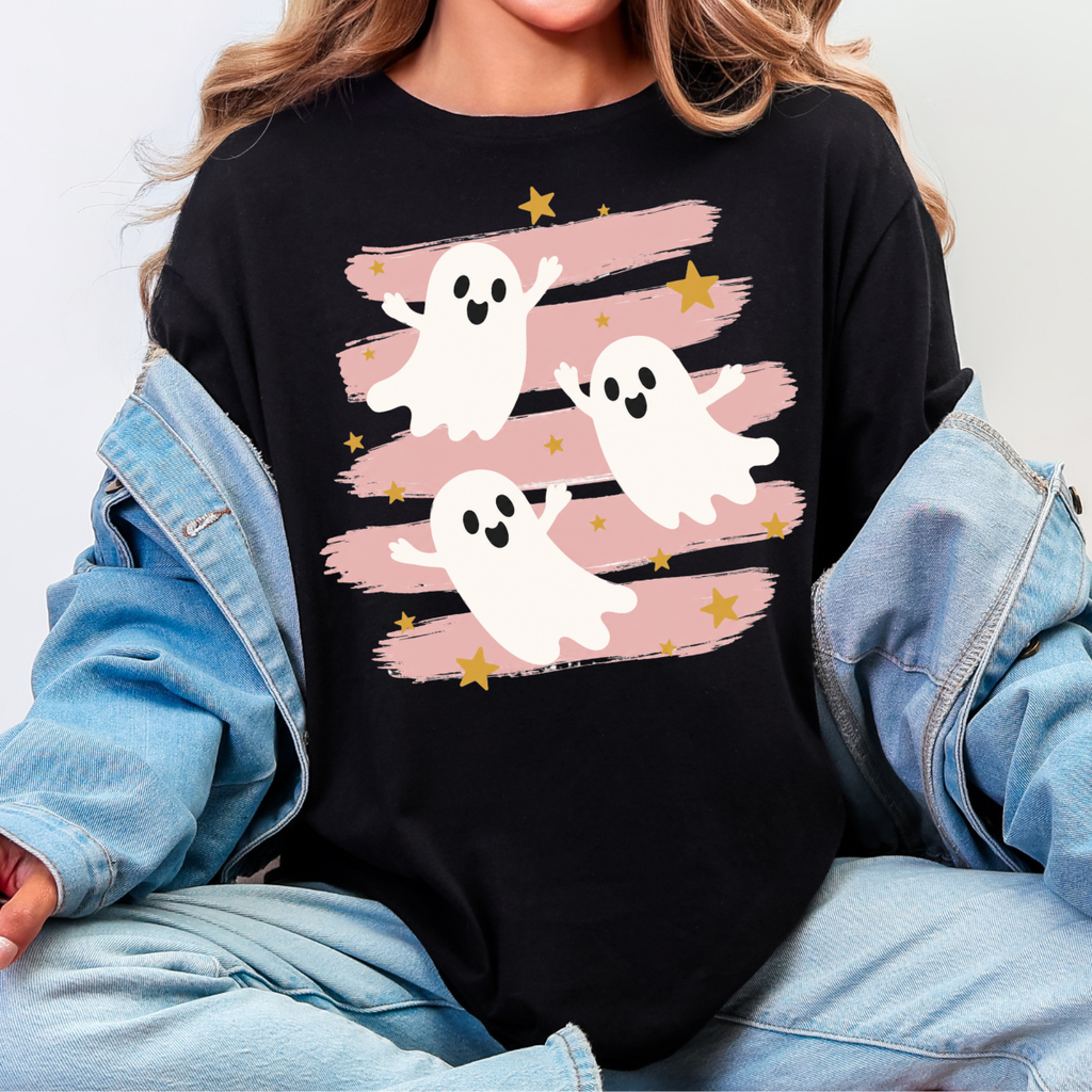 Woman wearing a black Halloween graphic tee with three white ghosts, pink brushstrokes, and gold stars.