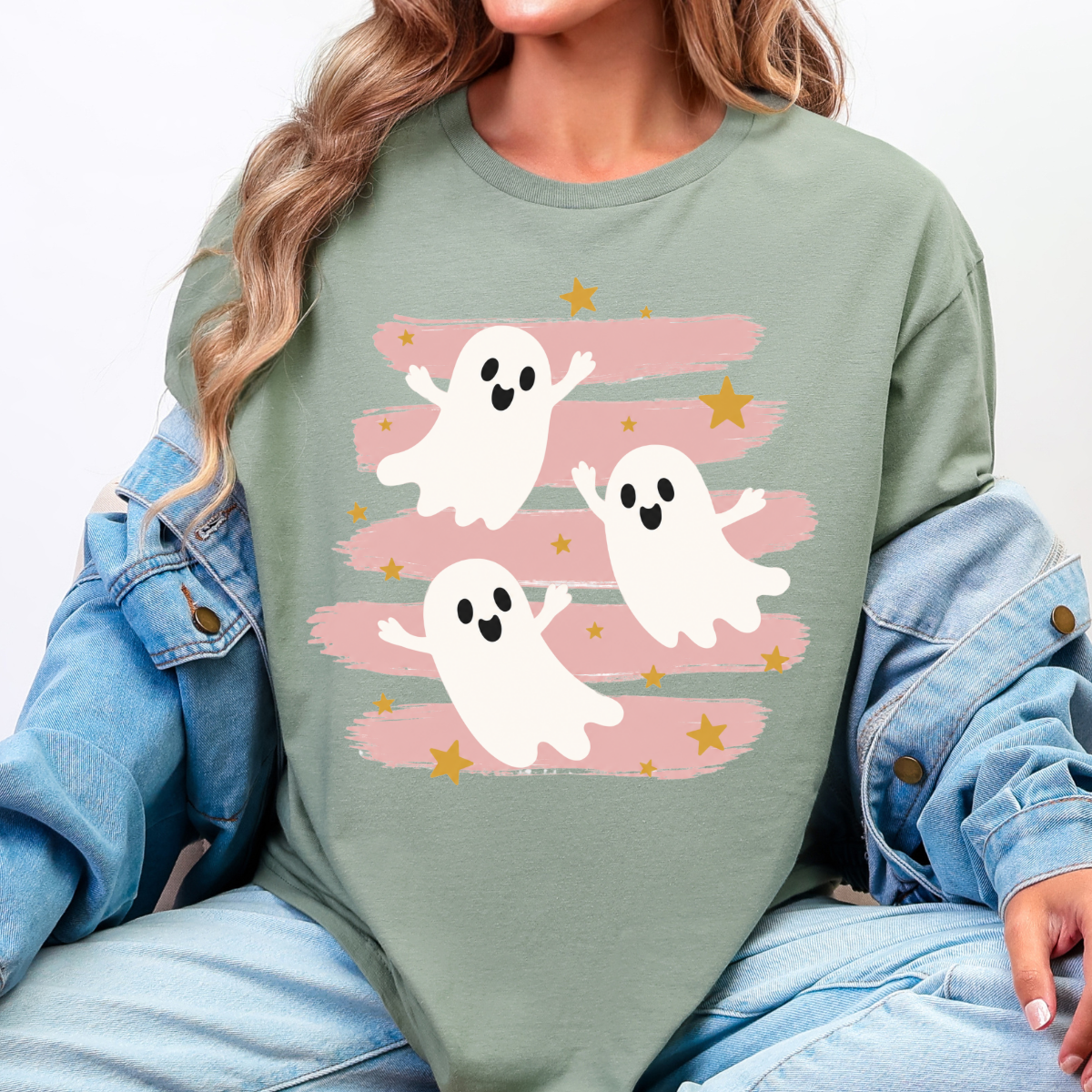 Woman wearing a sage green Halloween graphic tee with three white ghosts, pink brushstrokes, and gold stars.