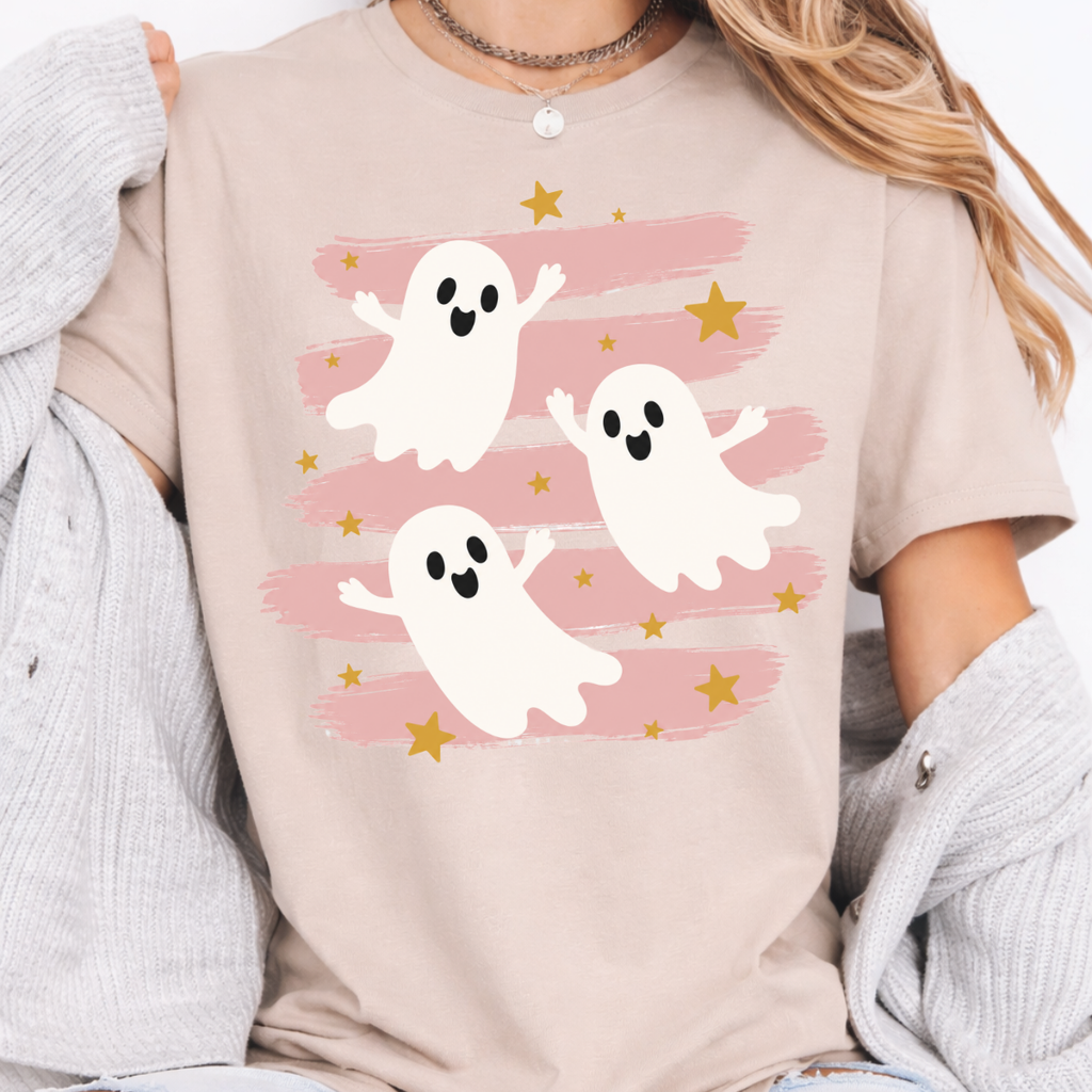 Woman wearing a sand Halloween graphic tee with three white ghosts, pink brushstrokes, and gold stars.