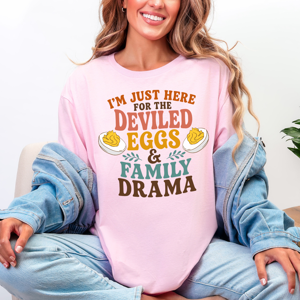Woman wearing a light pink Deviled Eggs and Family Drama Thanksgiving graphic tee with colorful lettering and deviled egg illustrations.