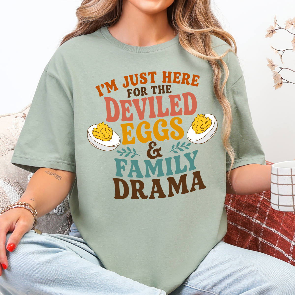 Woman wearing a sage Deviled Eggs and Family Drama Thanksgiving graphic tee with colorful lettering and deviled egg illustrations.