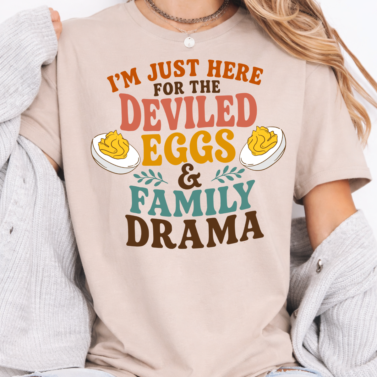 Woman wearing a sand Deviled Eggs and Family Drama Thanksgiving graphic tee with colorful lettering and deviled egg illustrations.