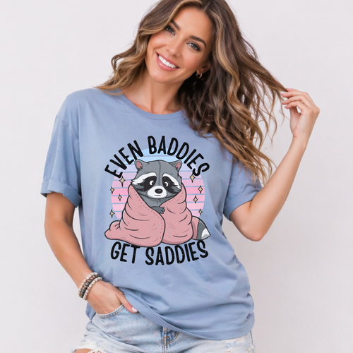 Woman wearing a blue t-shirt with a graphic of a raccoon and text that says "Even Baddies Get Saddies" on a plain background