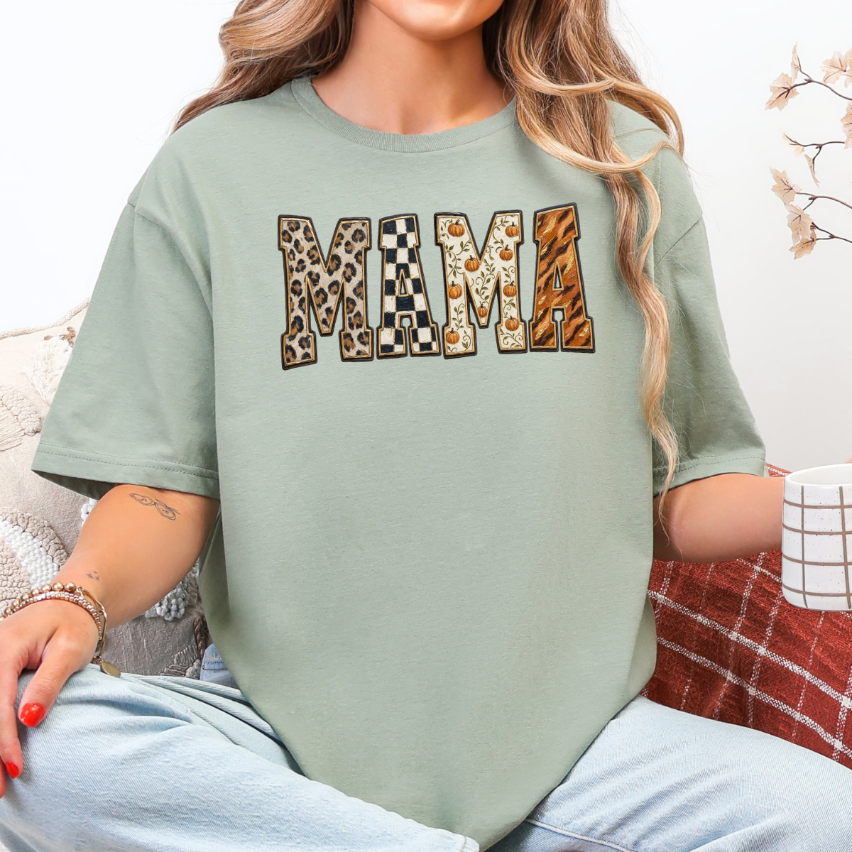 Woman wearing a sage Fall Mama graphic tee with leopard print, checkerboard, pumpkin, and orange patterned lettering.