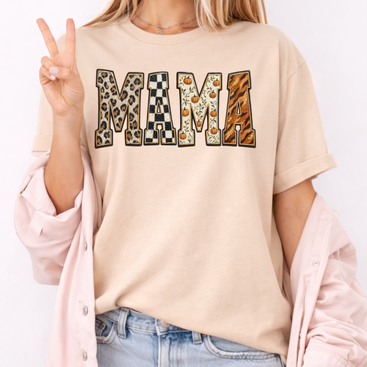 Woman wearing a whitesand Fall Mama graphic tee with leopard print, checkerboard, pumpkin, and orange patterned lettering.