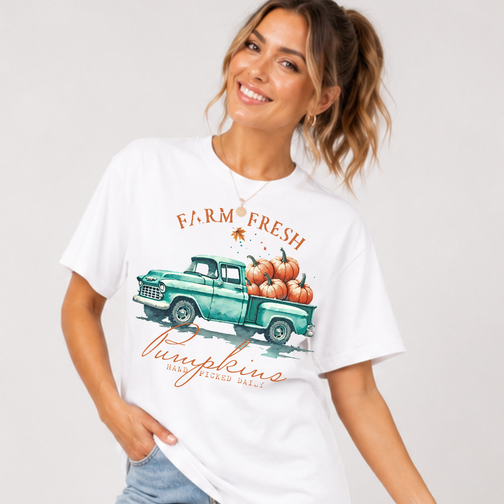 Woman wearing a white fall graphic tee with a teal vintage truck filled with orange pumpkins.