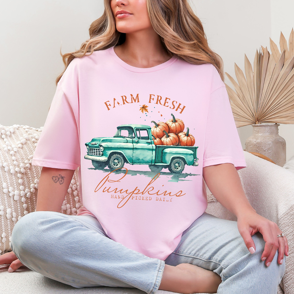 Woman wearing a light pink fall graphic tee with a teal vintage truck filled with orange pumpkins.