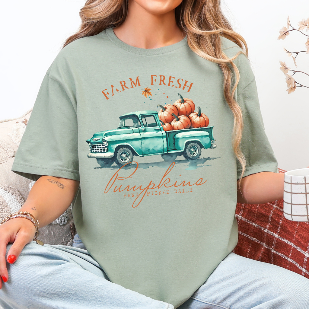 Woman wearing a sage fall graphic tee with a teal vintage truck filled with orange pumpkins.