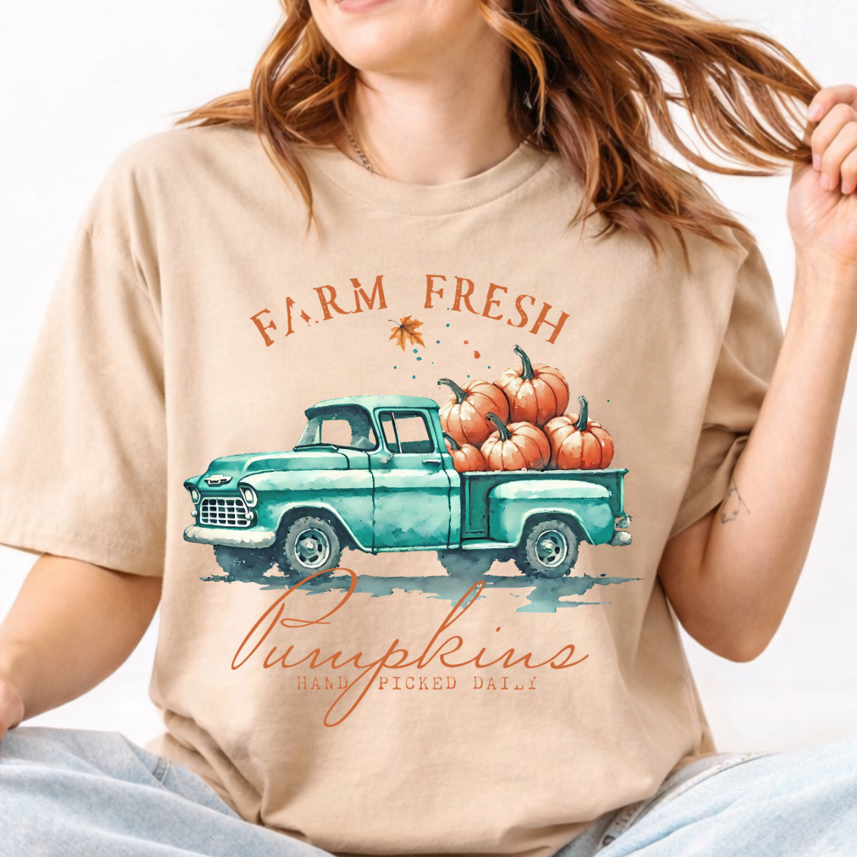 Woman wearing a sand fall graphic tee with a teal vintage truck filled with orange pumpkins.