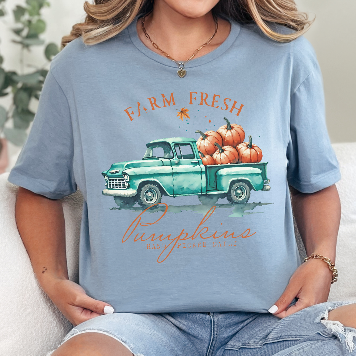 Woman wearing a stone blue fall graphic tee with a teal vintage truck filled with orange pumpkins.