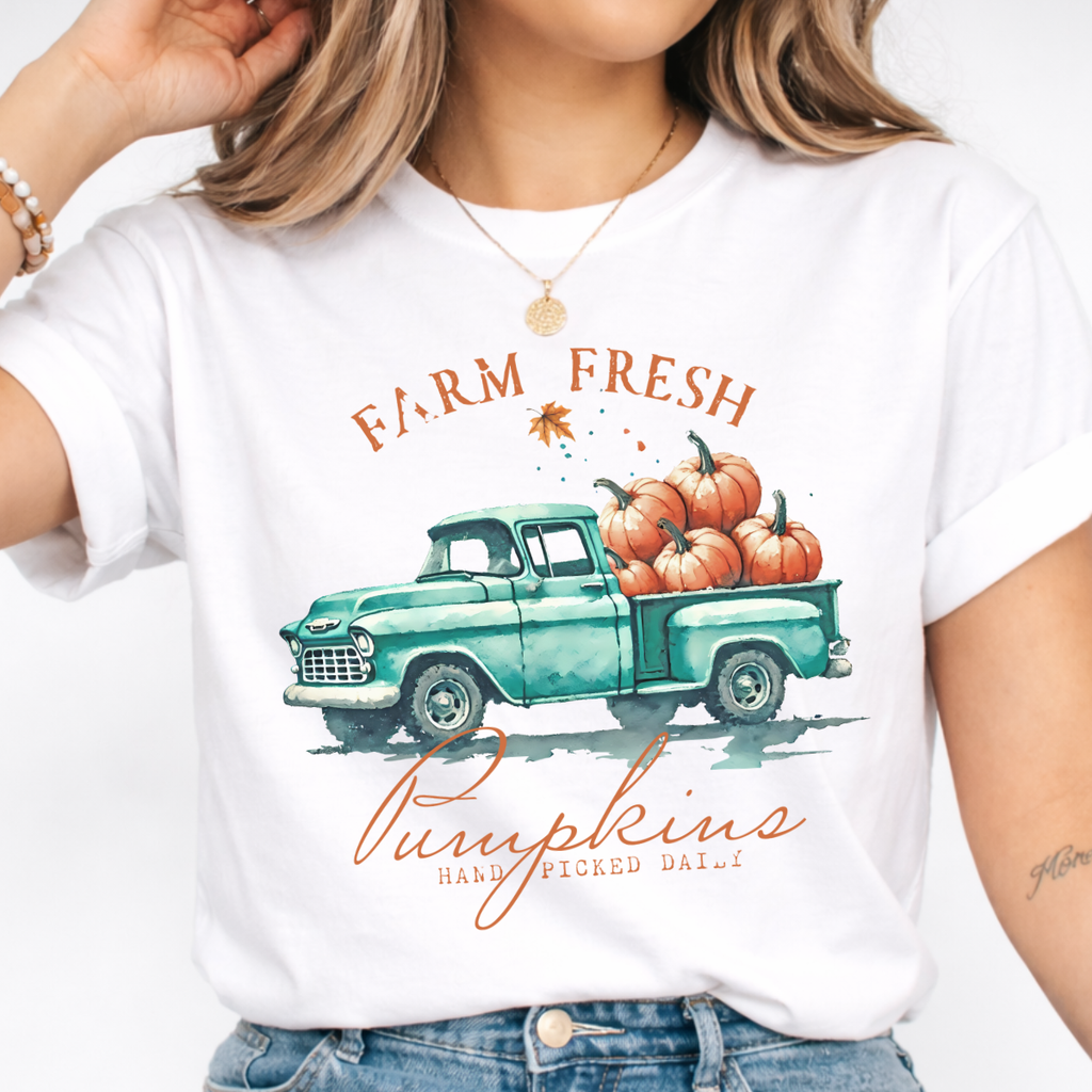 Woman wearing a white fall graphic tee with a teal vintage truck filled with orange pumpkins.