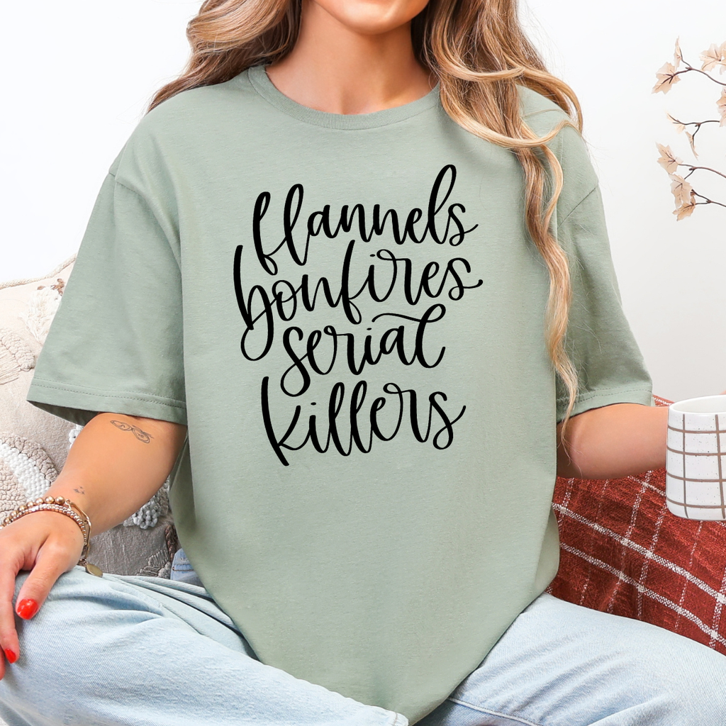 Woman wearing a sage green Flannels Bonfires and Serial Killers fall graphic tee with black script lettering.