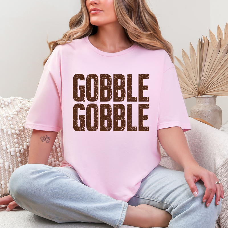 Woman wearing a light pink Gobble Gobble Thanksgiving graphic tee with distressed brown lettering.