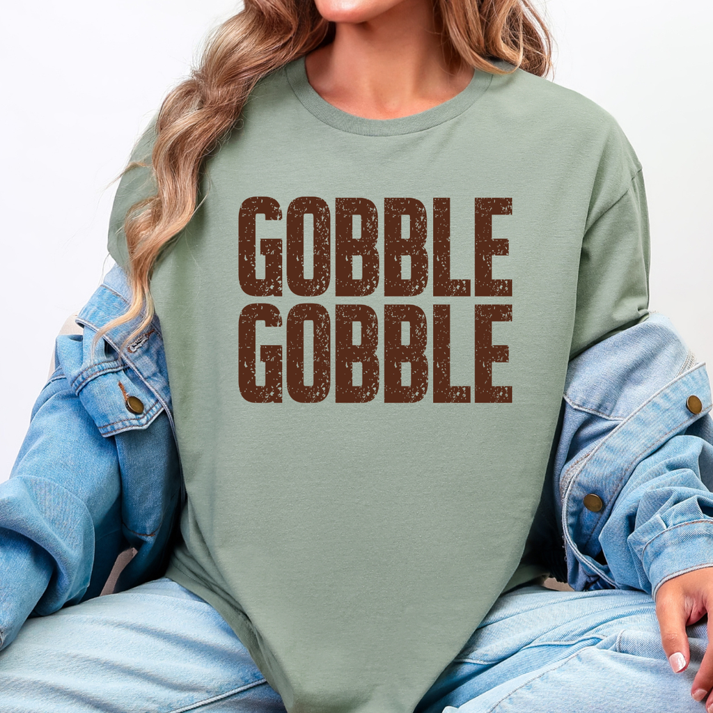 Woman wearing a sage Gobble Gobble Thanksgiving graphic tee with distressed brown lettering.