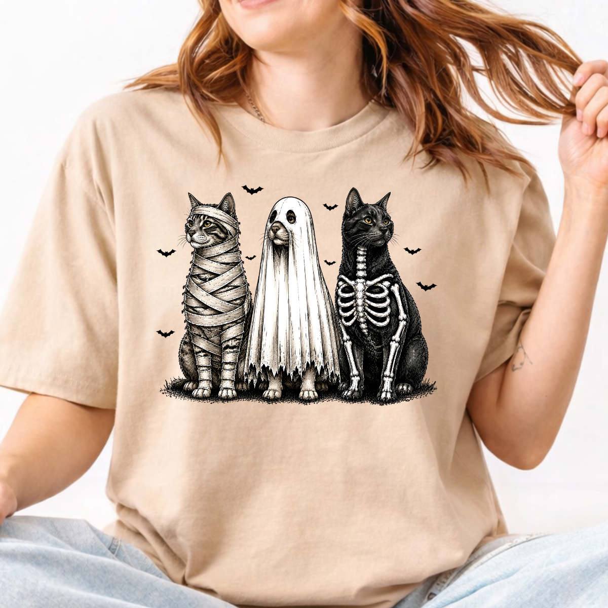 Woman wearing a sand Halloween cats graphic tee with mummy, ghost, and skeleton cats