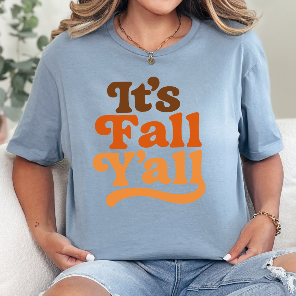 Woman wearing a stone blue It’s Fall Y’all graphic tee with brown and orange retro lettering.