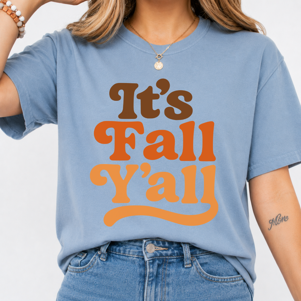 Woman wearing a stone blue It’s Fall Y’all graphic tee with brown and orange retro lettering.