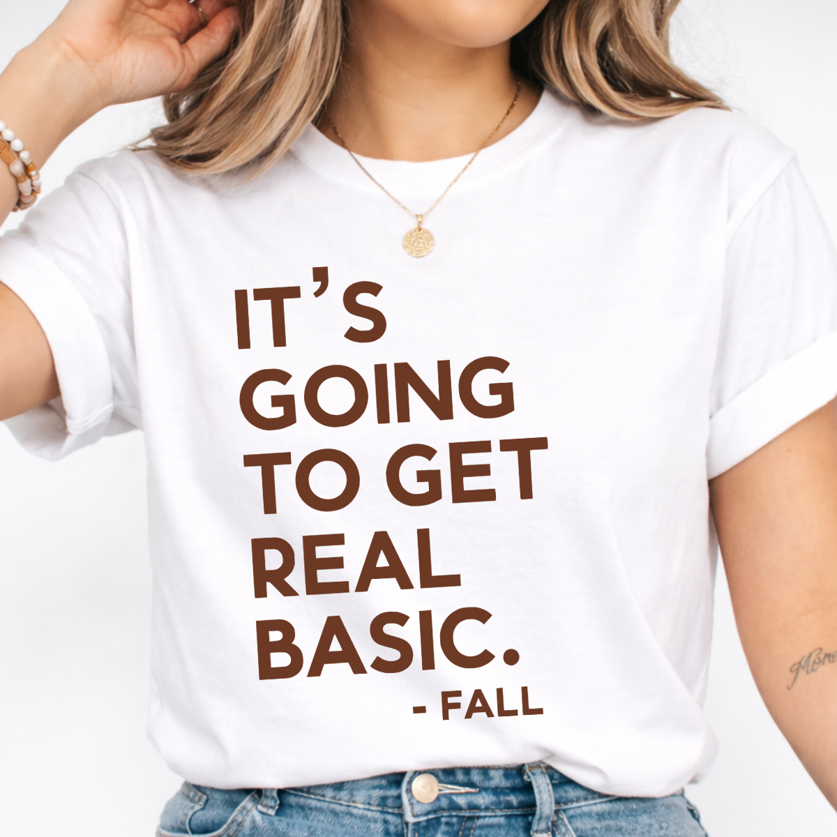 Woman wearing a white It’s Going to Get Real Basic fall graphic tee with bold brown lettering.