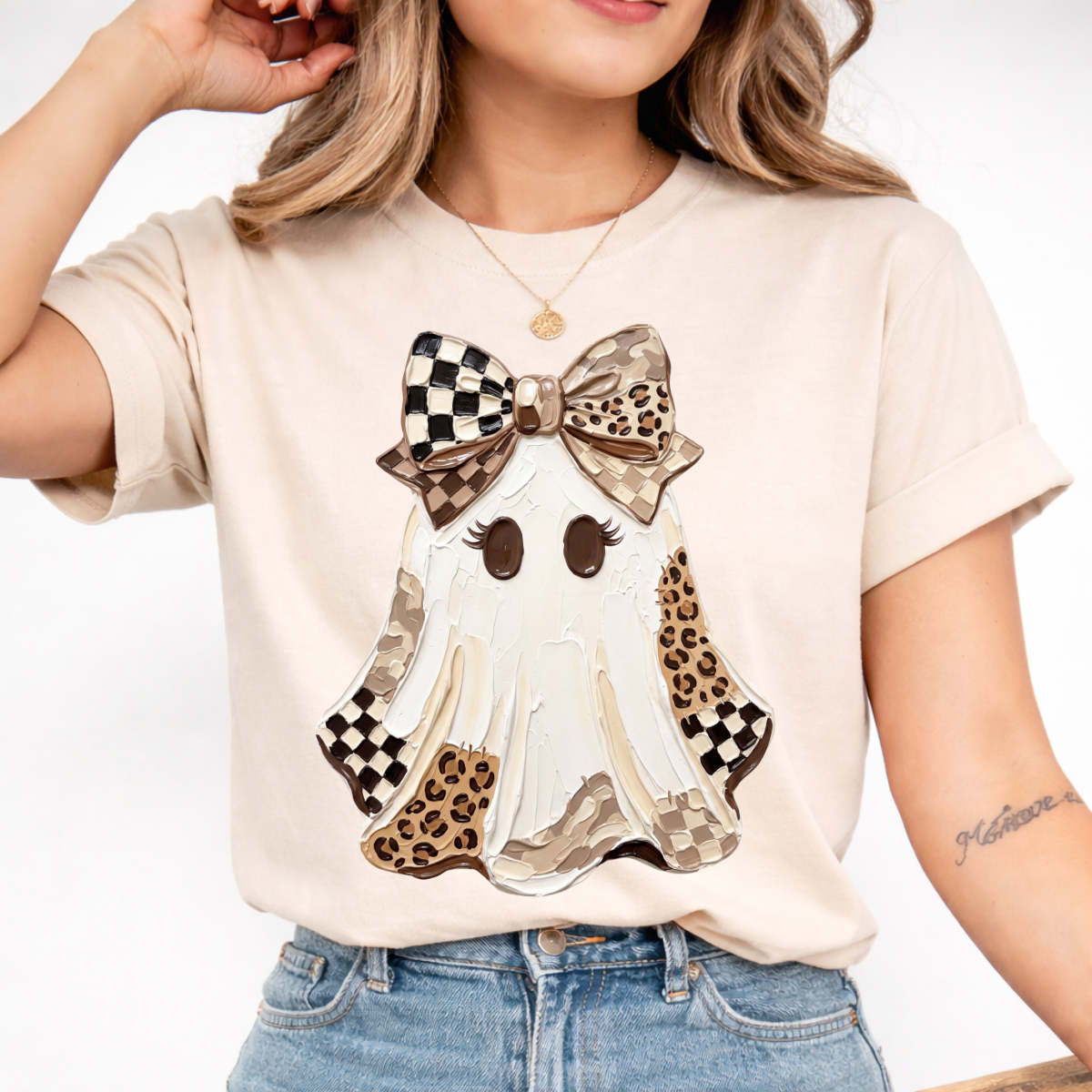 Woman wearing a sand Halloween graphic tee with a white ghost, leopard print and checkered details, and a large bow.