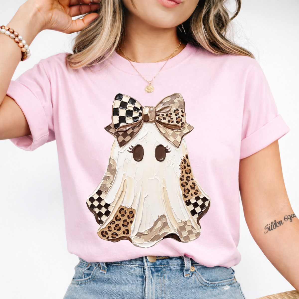 Woman wearing a light pink Halloween graphic tee with a white ghost, leopard print and checkered details, and a large bow.