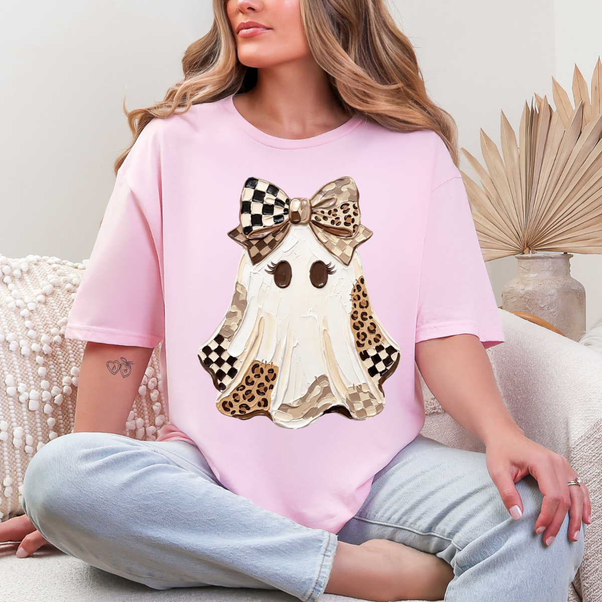 Woman wearing a light pink Halloween graphic tee with a white ghost, leopard print and checkered details, and a large bow.
