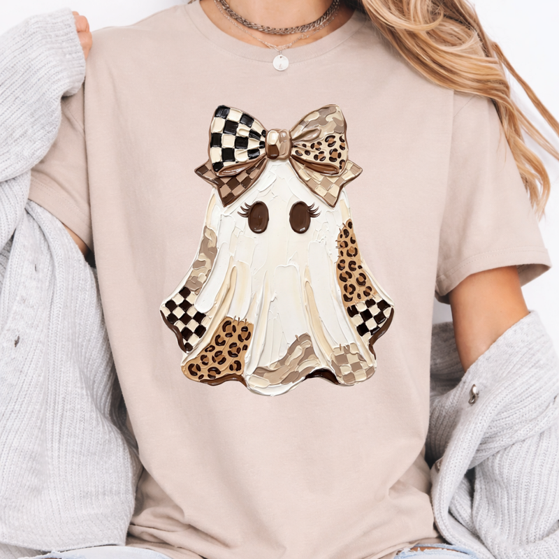 Woman wearing a sand Halloween graphic tee with a white ghost, leopard print and checkered details, and a large bow.