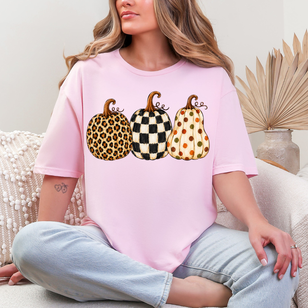 Woman wearing a light pink fall graphic tee with leopard, black-and-white checkered, and polka-dot pumpkins
