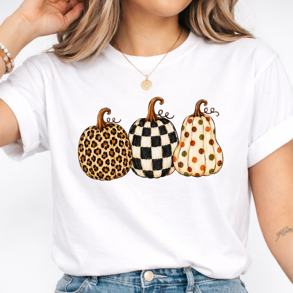 Woman wearing a white fall graphic tee with leopard, black-and-white checkered, and polka-dot pumpkins