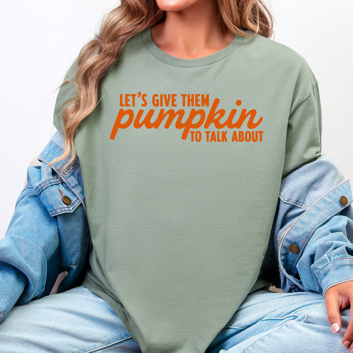 Woman wearing a sage Let’s Give Them Pumpkin to Talk About fall graphic tee with orange lettering.