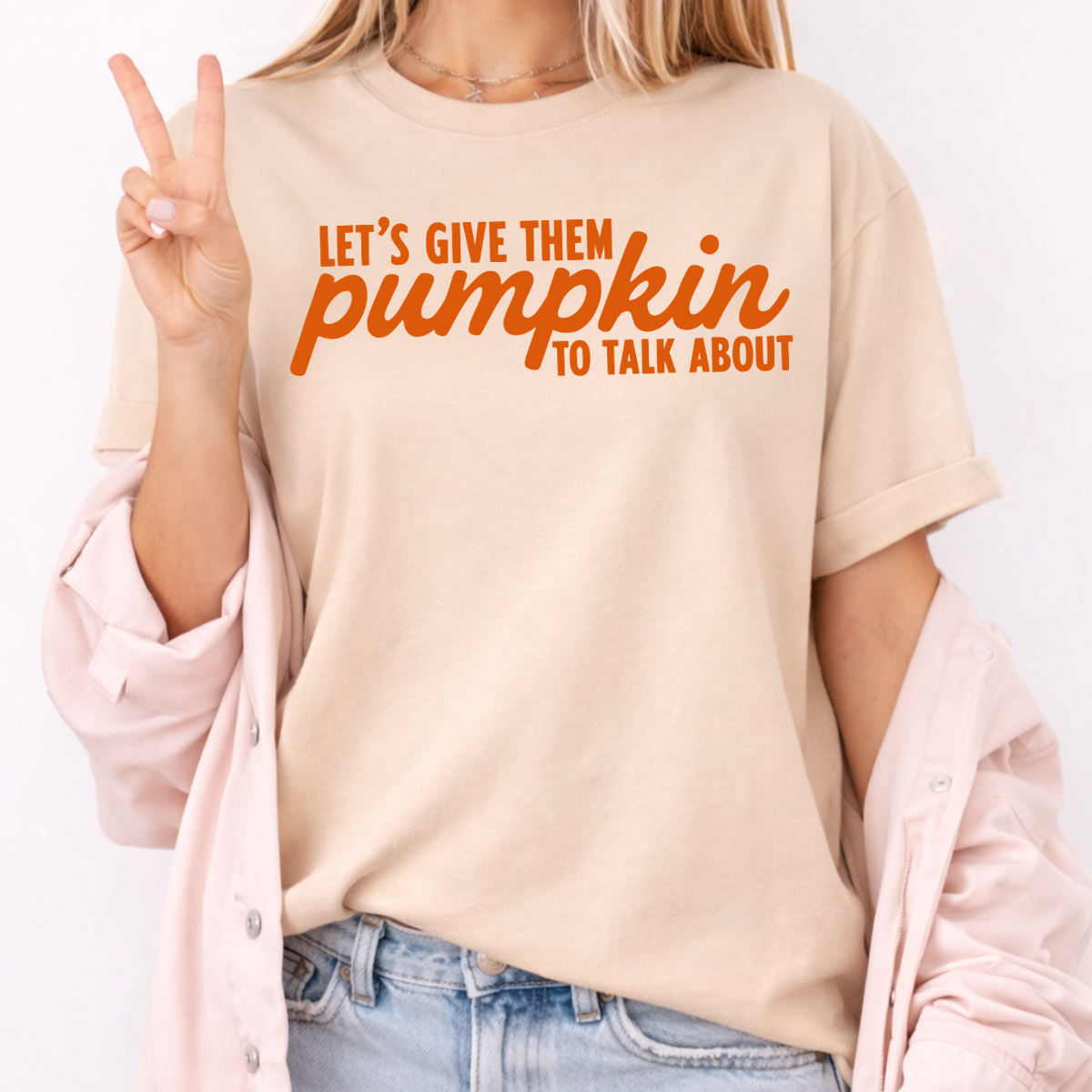 Woman wearing a sand Let’s Give Them Pumpkin to Talk About fall graphic tee with orange lettering.