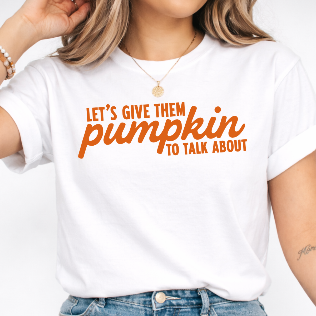 Woman wearing a white Let’s Give Them Pumpkin to Talk About fall graphic tee with orange lettering.