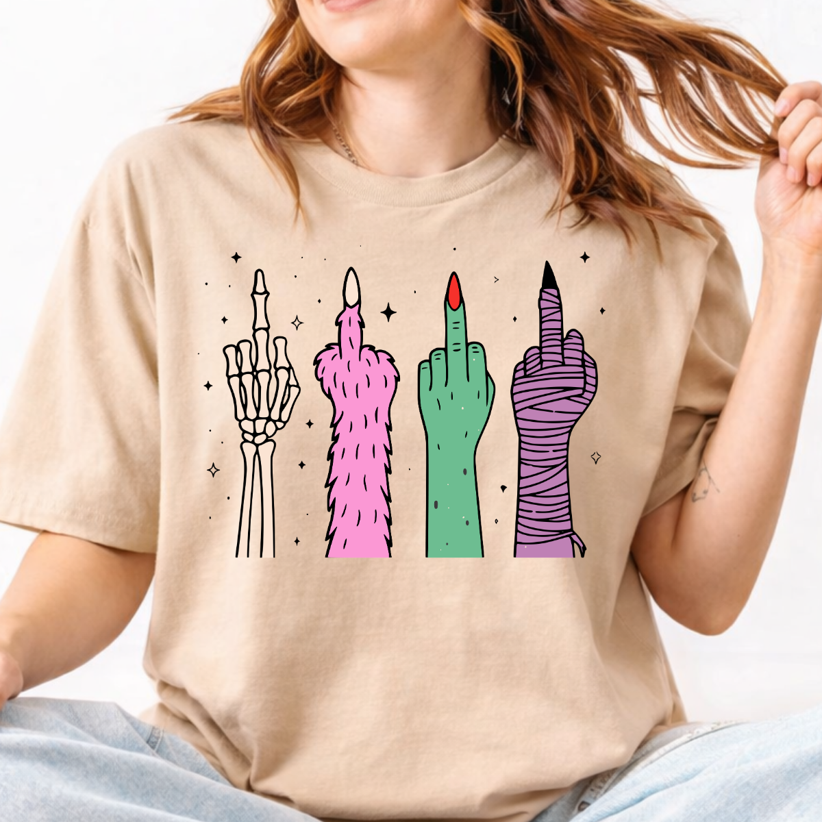 Woman wearing a sand Halloween graphic tee with colorful skeleton, monster, witch, and mummy middle finger hands.