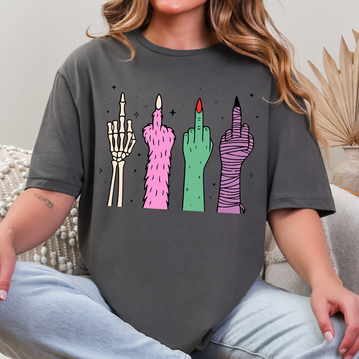 Woman wearing a charcoal Halloween graphic tee with colorful skeleton, monster, witch, and mummy middle finger hands.