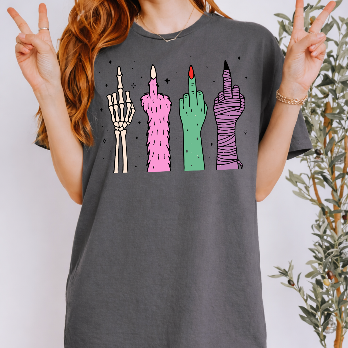 Woman wearing a charcoal Halloween graphic tee with colorful skeleton, monster, witch, and mummy middle finger hands.
