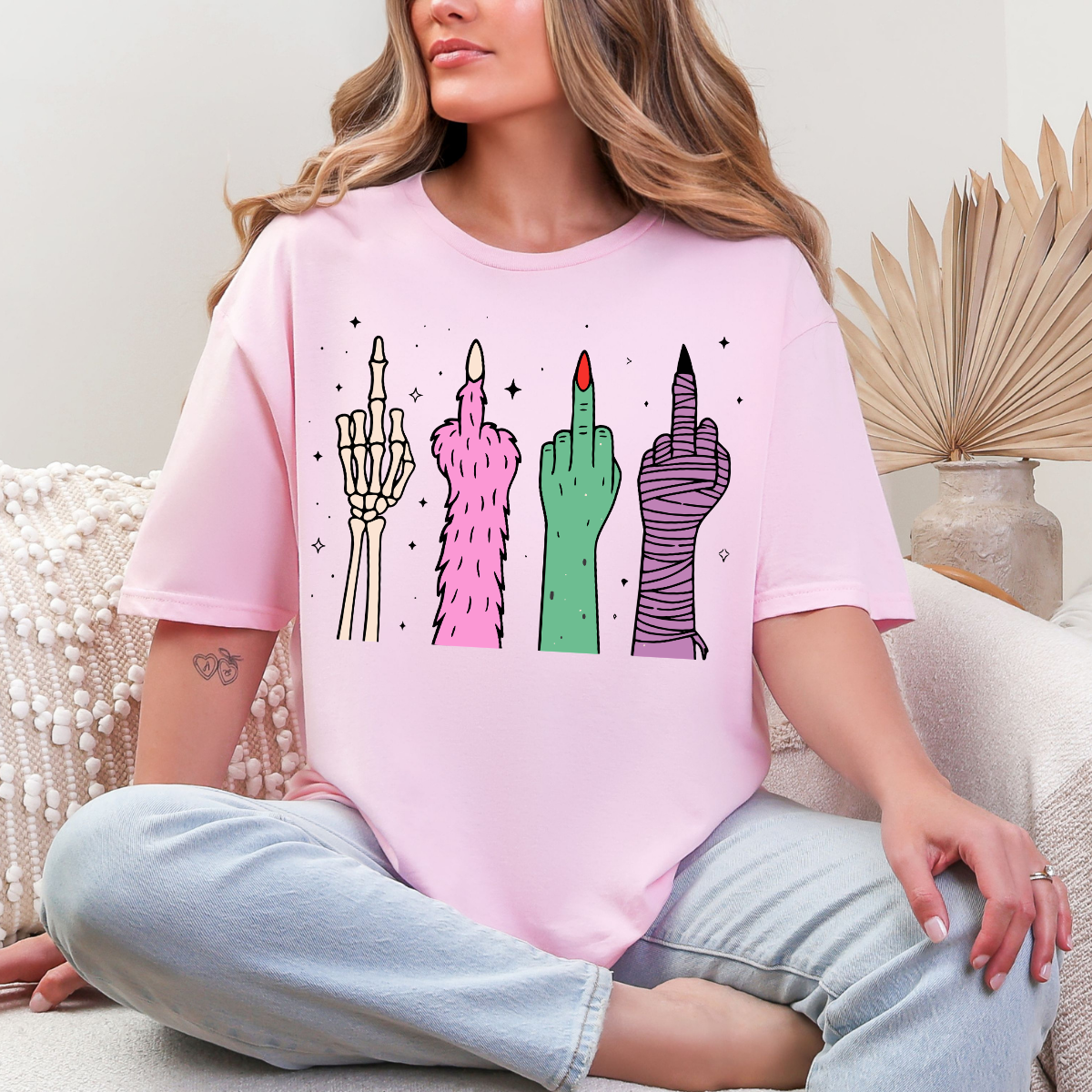 Woman wearing a light pink Halloween graphic tee with colorful skeleton, monster, witch, and mummy middle finger hands.