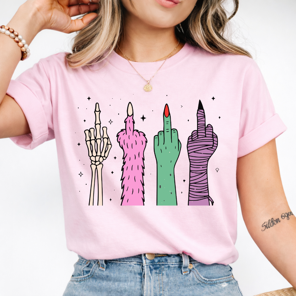 Woman wearing a light pink Halloween graphic tee with colorful skeleton, monster, witch, and mummy middle finger hands.