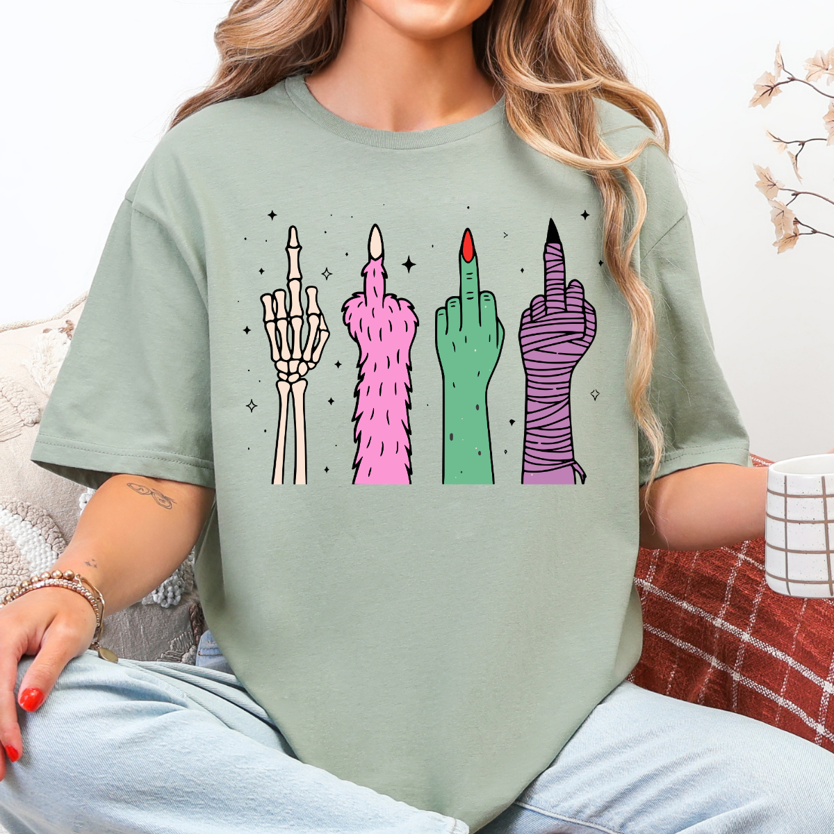 Woman wearing a sage Halloween graphic tee with colorful skeleton, monster, witch, and mummy middle finger hands.