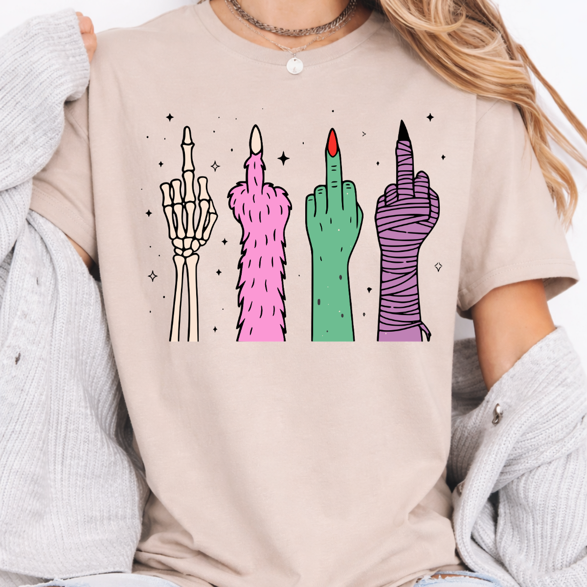 Woman wearing a sand Halloween graphic tee with colorful skeleton, monster, witch, and mummy middle finger hands.