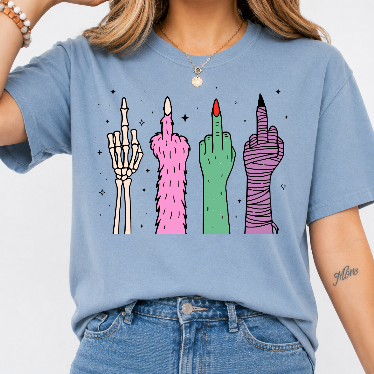 Woman wearing a stone blue Halloween graphic tee with colorful skeleton, monster, witch, and mummy middle finger hands.