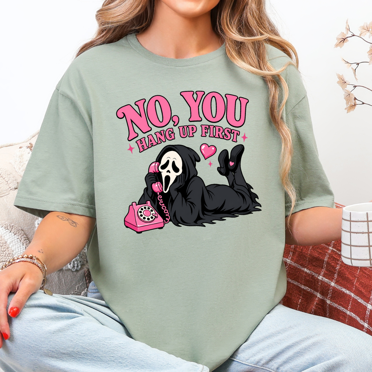 Woman wearing a sage No You Hang Up First Halloween graphic tee with a masked horror character and pink telephone.