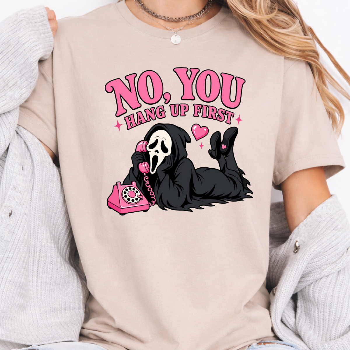 Woman wearing a sand No You Hang Up First Halloween graphic tee with a masked horror character and pink telephone.